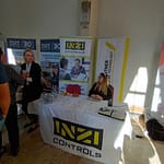 Kiállítói stand beltérben, asztalnál ülő és álló résztvevőkkel, roll-up molinókkal.