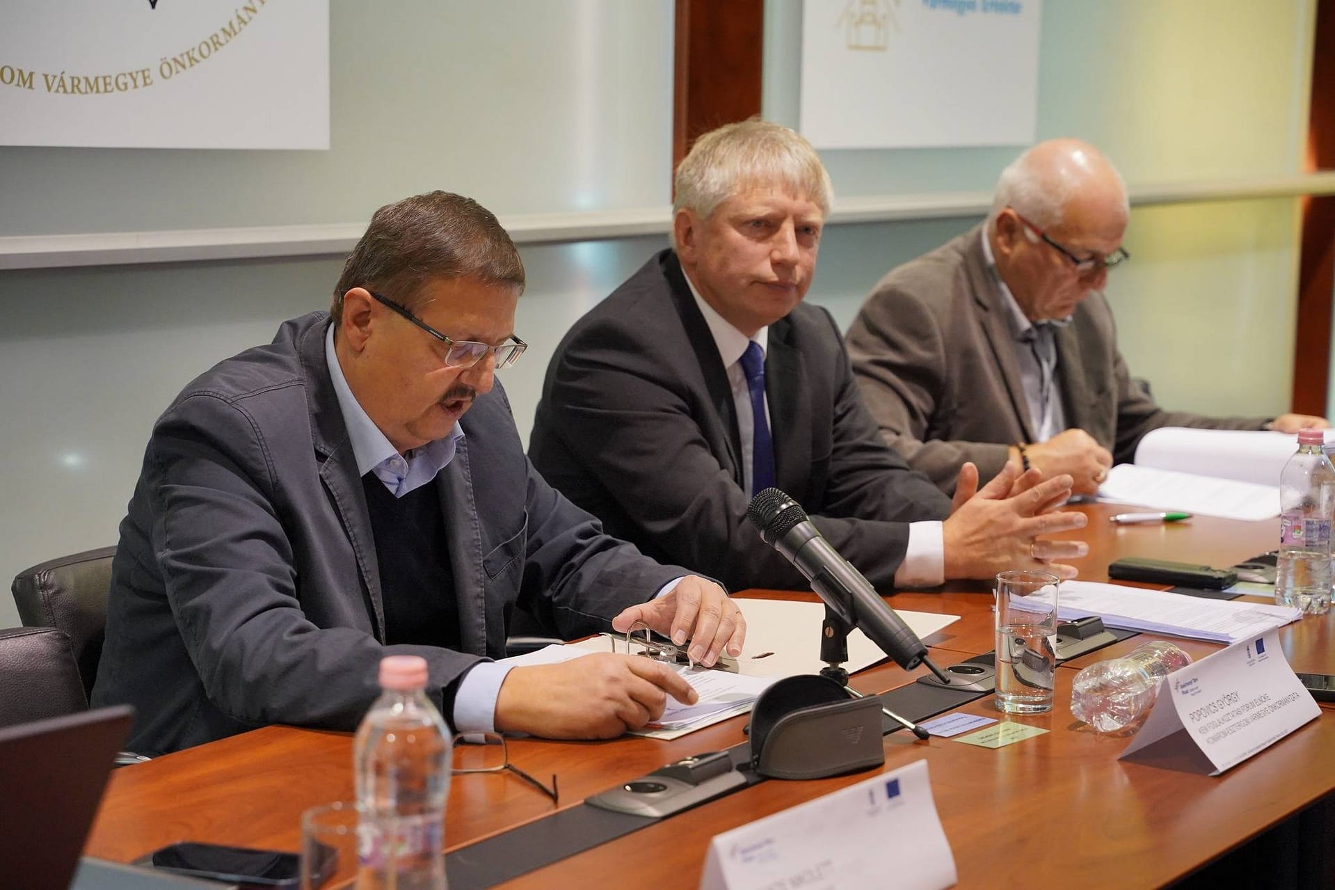 Popovics György elnök, Kancz Csaba főispán, Borsó Tibor alelnök szakmai döntéshozók beszélgetnek egy tanácskozáson, előttük dokumentumok és mikrofonok.