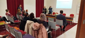 Résztvevők ülnek a teremben, elöl előadók állnak.