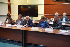 Munkaértekezlet résztvevői ülnek egy hosszú tárgyalóasztalnál, előttük névtáblák és iratok.