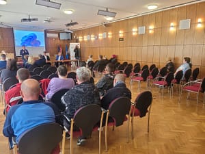 Előadás egy közösségi teremben, a hallgatóság sorokban ül és a kivetített prezentációt követi.